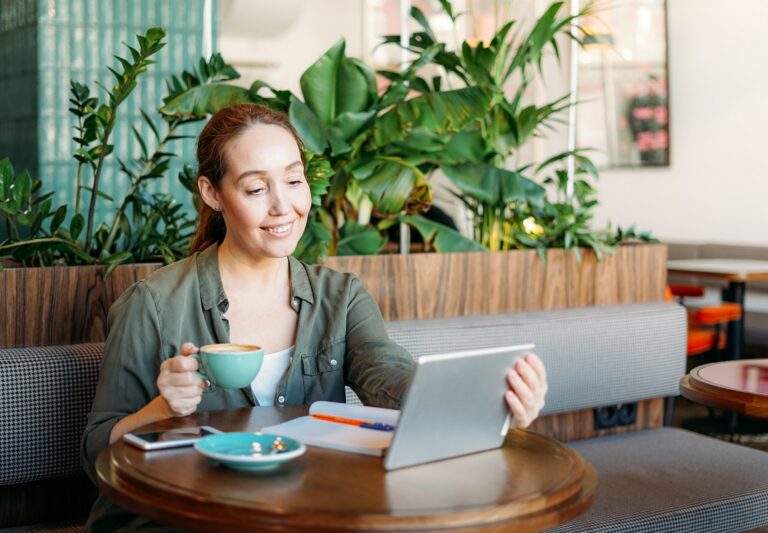 Adult smiling brunette woman doing notes and look at tablet takes online course on table at cafe